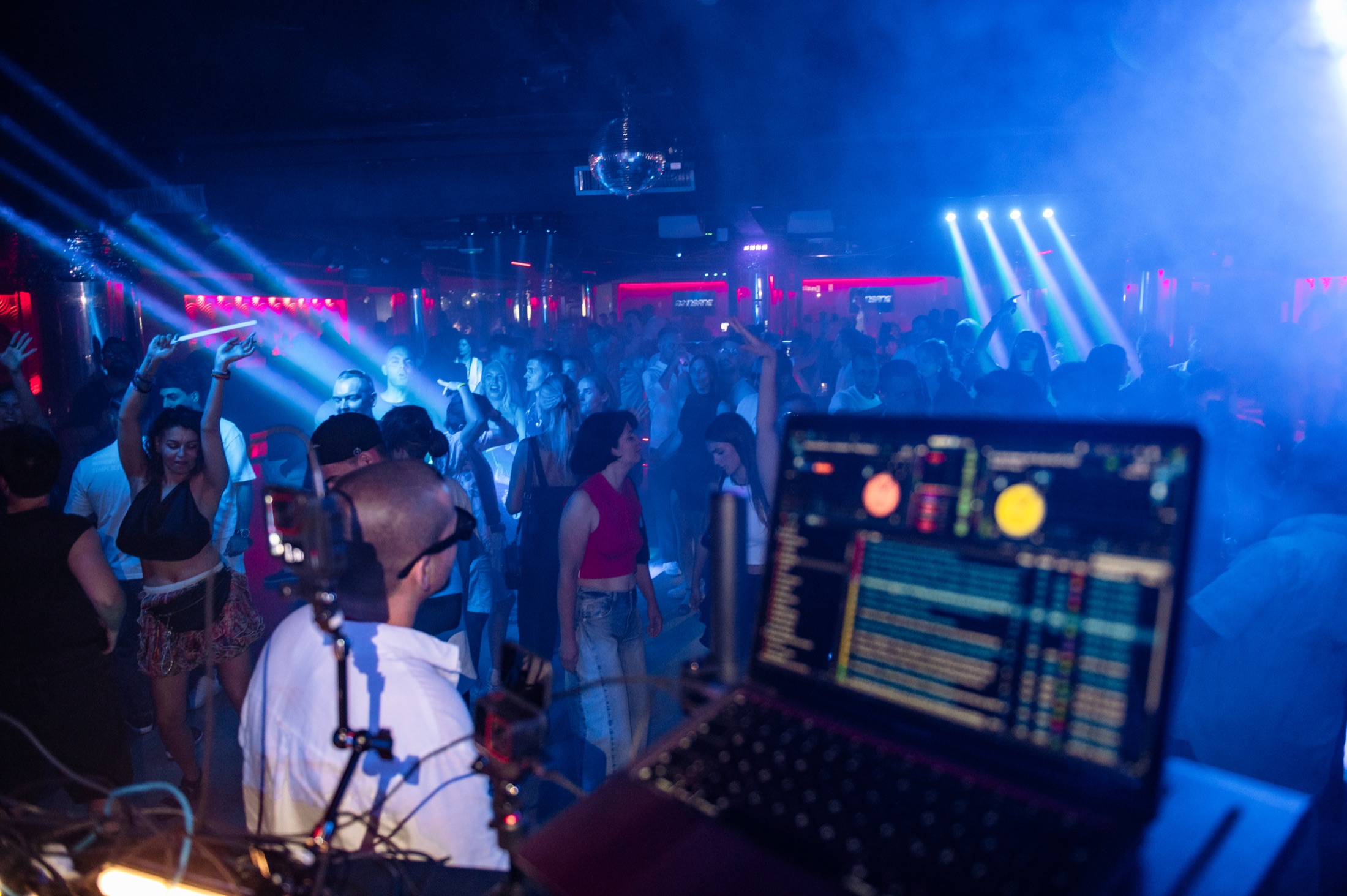 Packed dance floor seen from the DJ booth under blue lights at a [nɛbó:] night
